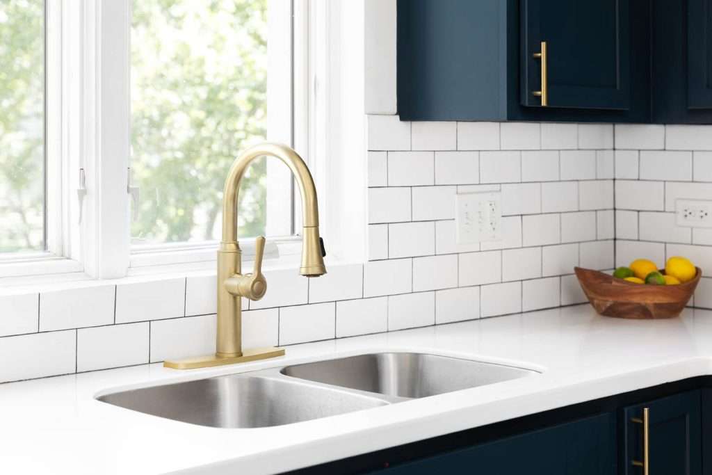 A kitchen detail with a gold faucet in front of a window, white subway tile backsplash, a bowl of fruit on the white marble countertop, and blue cabinets.